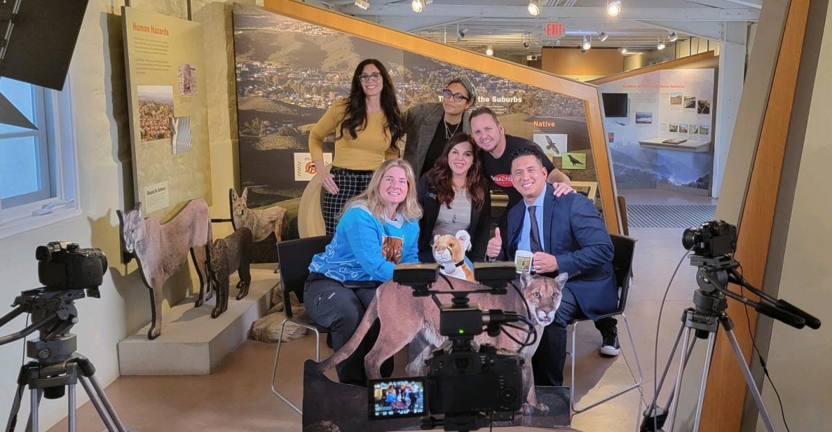 A group of men and women posing for a picture next to podcasting equipment and cardboard cutouts of wildlife.