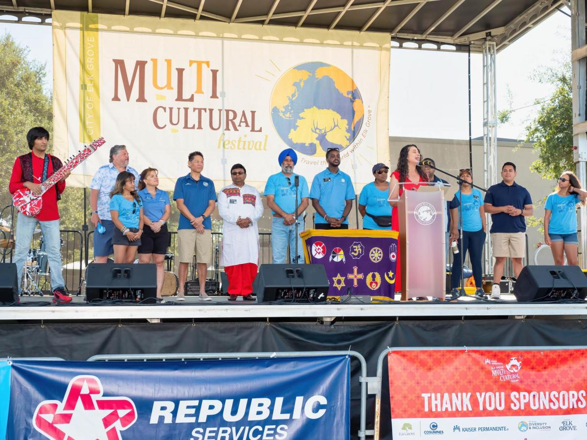 A mayor speaking on stage during a festival, flanked by a diverse group of residents.
